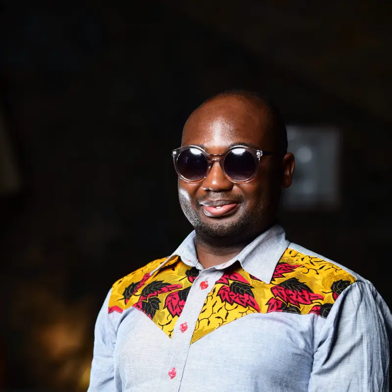 Theophilus Michael Odaudu, a black man wearing sunglasses and a white shirt with yellow and red patterns at the shoulders, smilling in front of a dark background