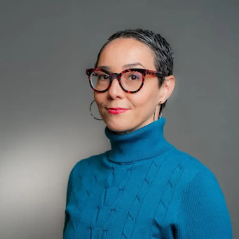 Violeta Rubiani, with short shaved black hair, wearing a blue turtleneck, red glasses and pink lipstick, looking into the camera on a grey background
