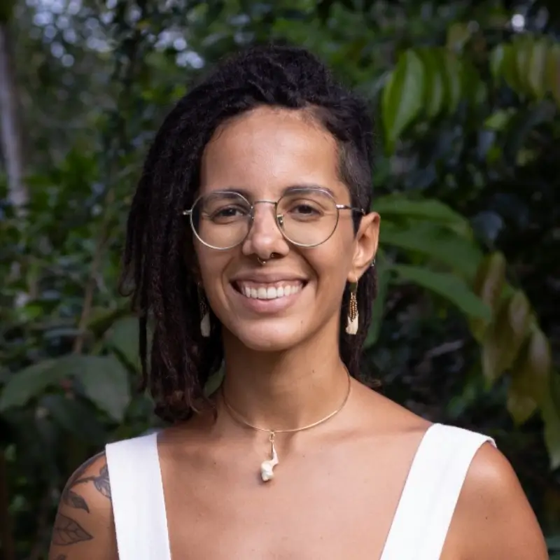 Verônica Veloso, with dreads, wearing a white shirt, thin prescription glasses, a silver necklace and gold earrings, smilling to the camera in front of greenery