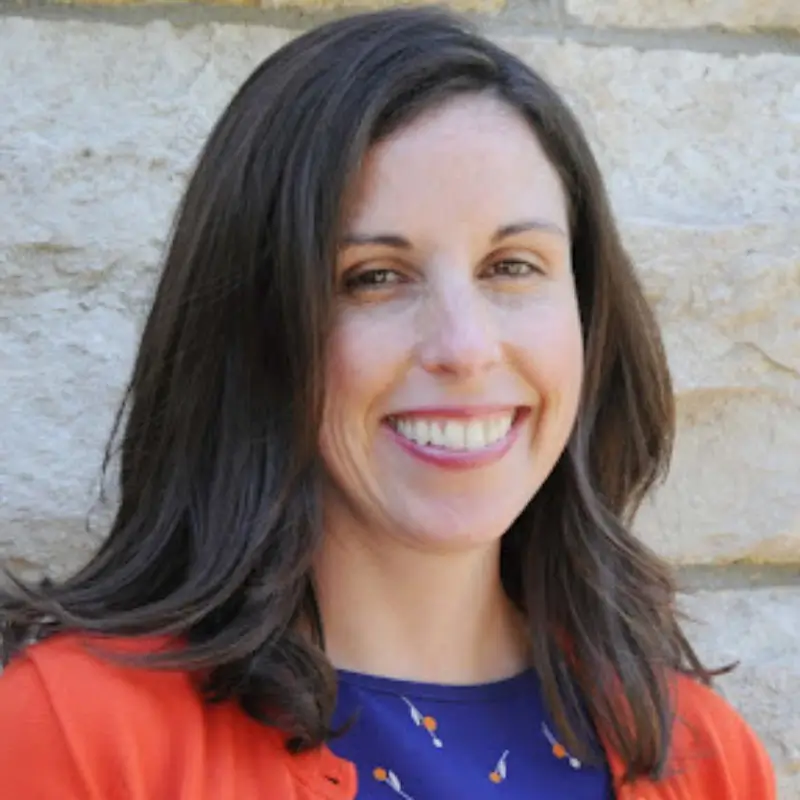 Eileen Horn, a white brunettem wearing a blue shirt and a orange cardigan, smilling into the camera in front of a gray stone wall.