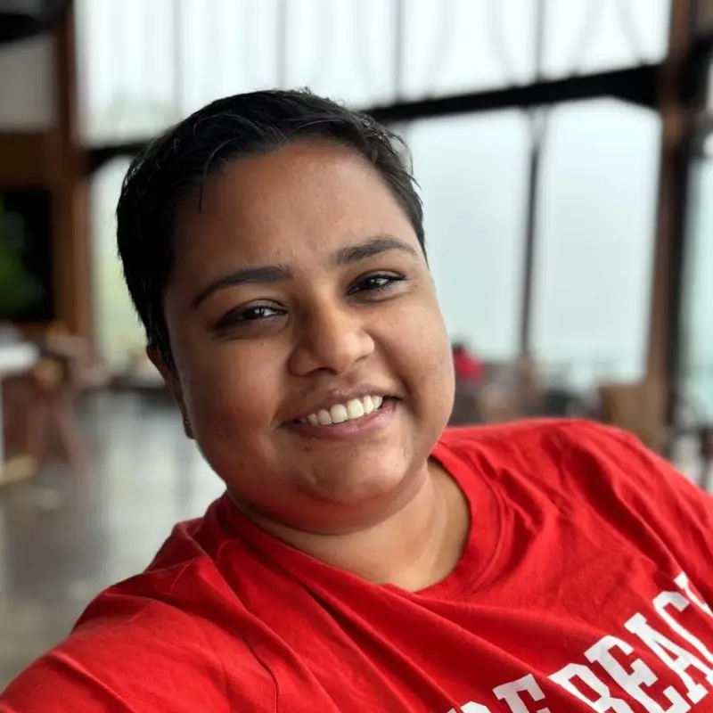 Anamika Dutt, with short dark hair, wearing a red shirt, smiling while seated indoors with large windows in the background.