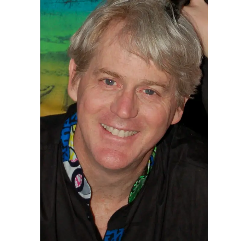 Chris Allan, a middle-aged man with light skin and gray hair smiles at the camera, wearing a black shirt with a colorful patterned collar.