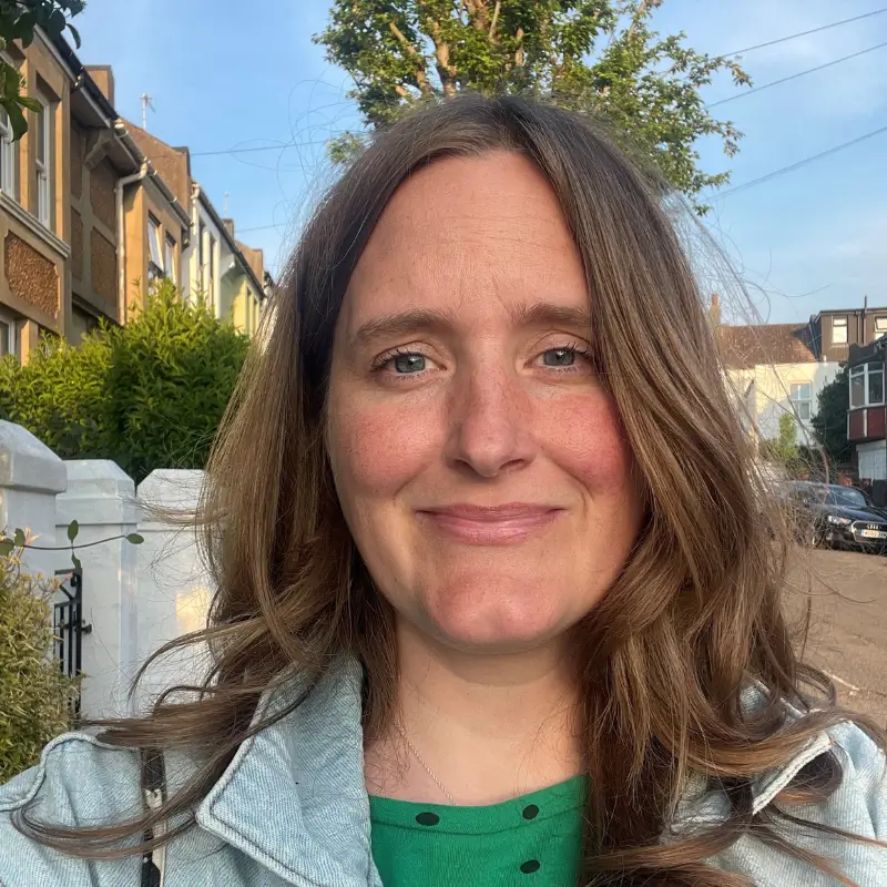 Zoe Corden, a white blonde, wearing a green shirt with a light blue denim jacket, smilling at the camera in front of a street on a sunny day