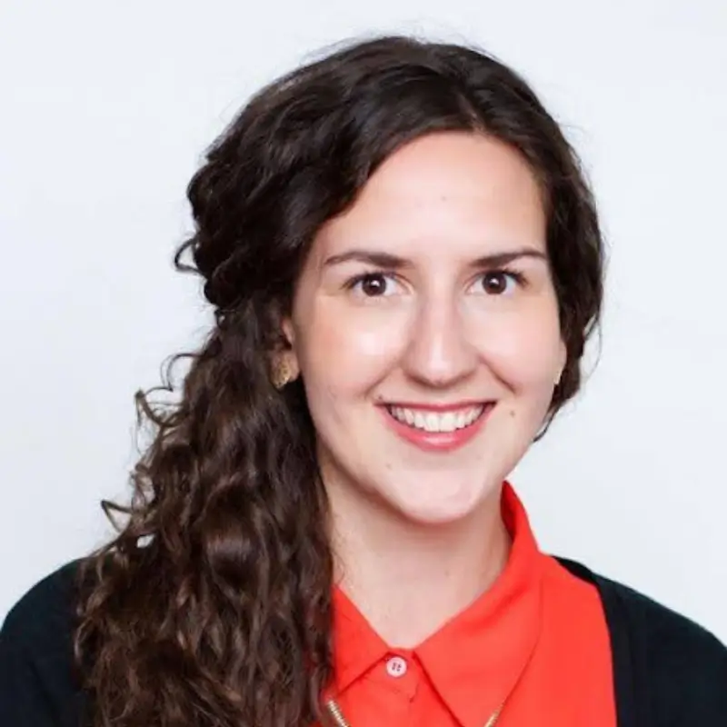 Caitlin O’Brien, a brunette with a red button up shirt and a black jacket, smilling to the camera in front of a white background