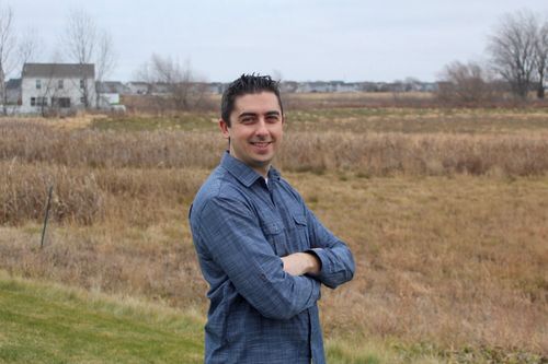 Christopher Pinski stands at an angle to the camera with arms folded, smiling. He wears a blue button-down shirt. In the background is a field with bare trees and a white house, pictured on a cloudy day.
