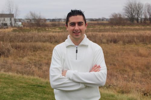 Christopher Pinski in white zip sweatshirt smiles with arms folded; it's a cloudy day, and behind him is a field with bare trees and a white house in the distance.