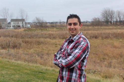 Christopher Pinski stands at an angle to the camera with arms folded, smiling. He wears a red, white, and black longsleeve flannel button-down shirt. He stands in a field with bare trees and a white house in the distance. It's a cloudy day.