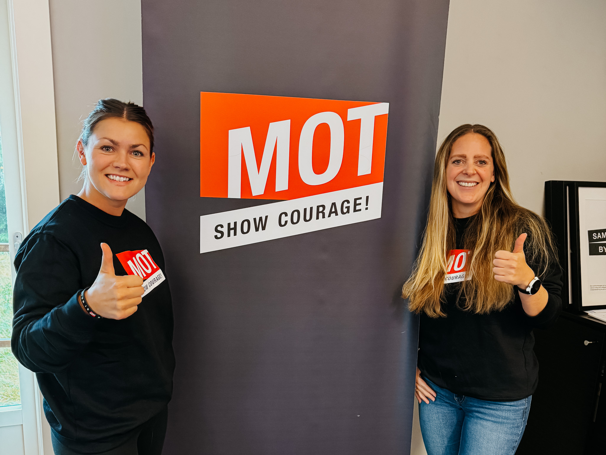 To MOT coacher fra Halsa Barne og ungdomsskole smiler og gir tommel opp ved MOT logoen under Grunnutdanning på MOT Camp