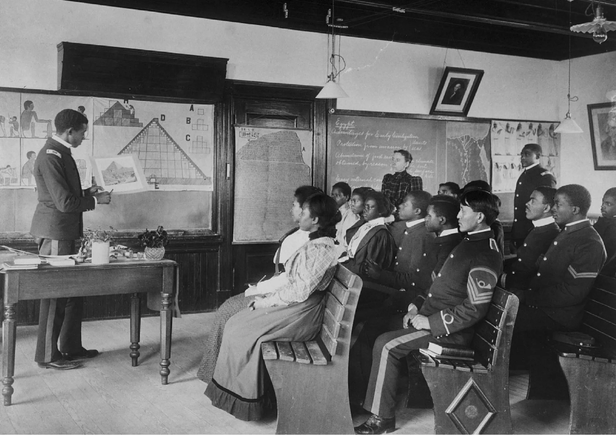 A historical black and white photograph of a classroom. A lecturer is standing in front of several rows of wooden benches filled with students of color, pointing at a picture that he is holding up. There are blackboards and posters on the classroom walls.