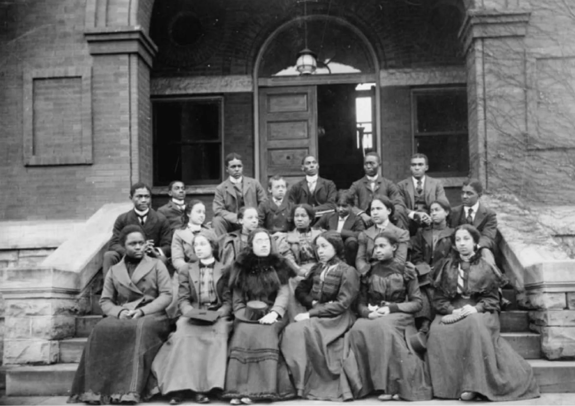 A black and white historical photograph of a group of smartly dressed Black students sitting on the stairs to the entrance of a large university building.