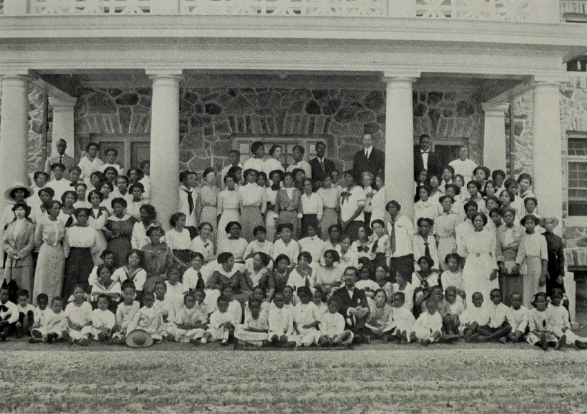 A historical sepia photo of a large group of smartly dressed Black adults and youth. They stand on the porch of a grand building, the Cheyney Training School for Teachers