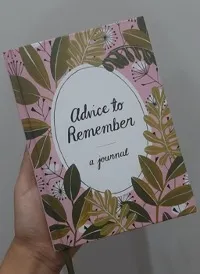 A hand is holding a book with a pink cover and illustrations of green leaves and ferns. The cover text reads, "Advice to Remember: A Journal."