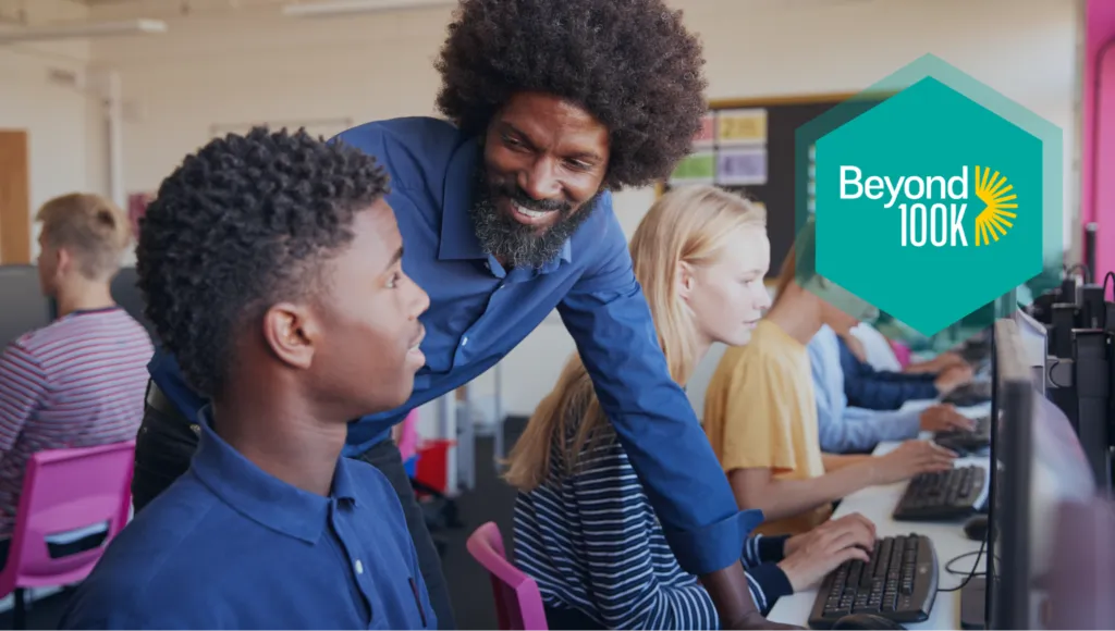 A Black teacher with dark, curly hair in an afro style smiles and assists a Black student at a computer. Other students work on computers in the background. A logo that says "Beyond 100K" is superimposed on the right side.