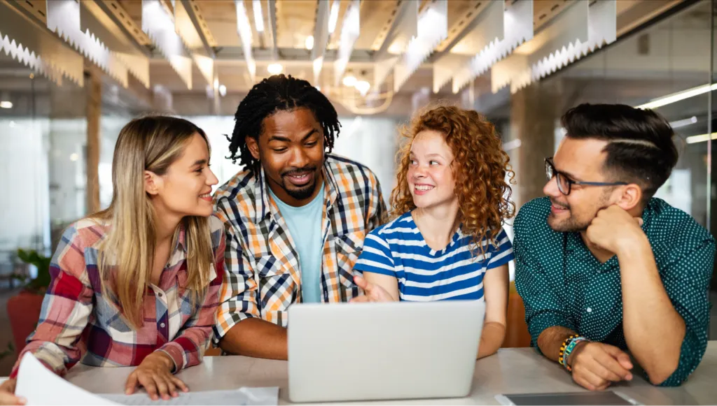 Four people of different races and genders sit at a table around an open laptop in a library. They are smiling at each other and look like they’re in deep conversation.