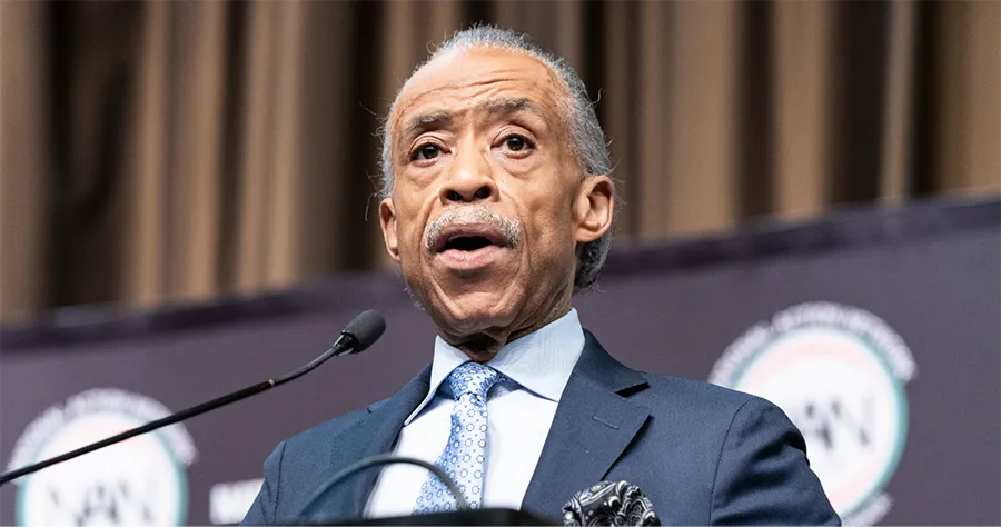 Reverend Al Sharpton, an elderly mixed-race man, speaking into a mic at a podium. He has short gray hair and a gray mustache. He wears a black suit jacket with a blue shirt and blue tie. He looks serious.