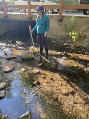 A woman stands in a shallow river next to a bridge culvert. She is holding a net.