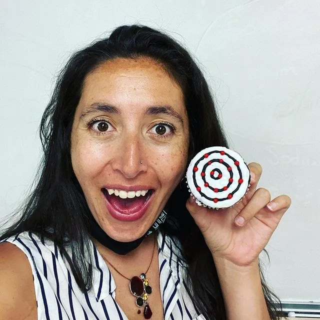 Picture of Autumn Rivera, a woman with dark hair wearing a black and white striped shirt and smiling broadly. She holds a cupcake up in her left hand that has decorations on top in the pattern of an element.