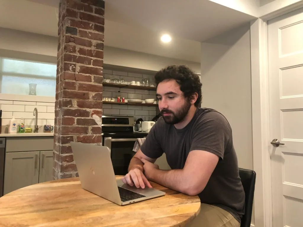 Bearded, white man seated at a table using a laptop.