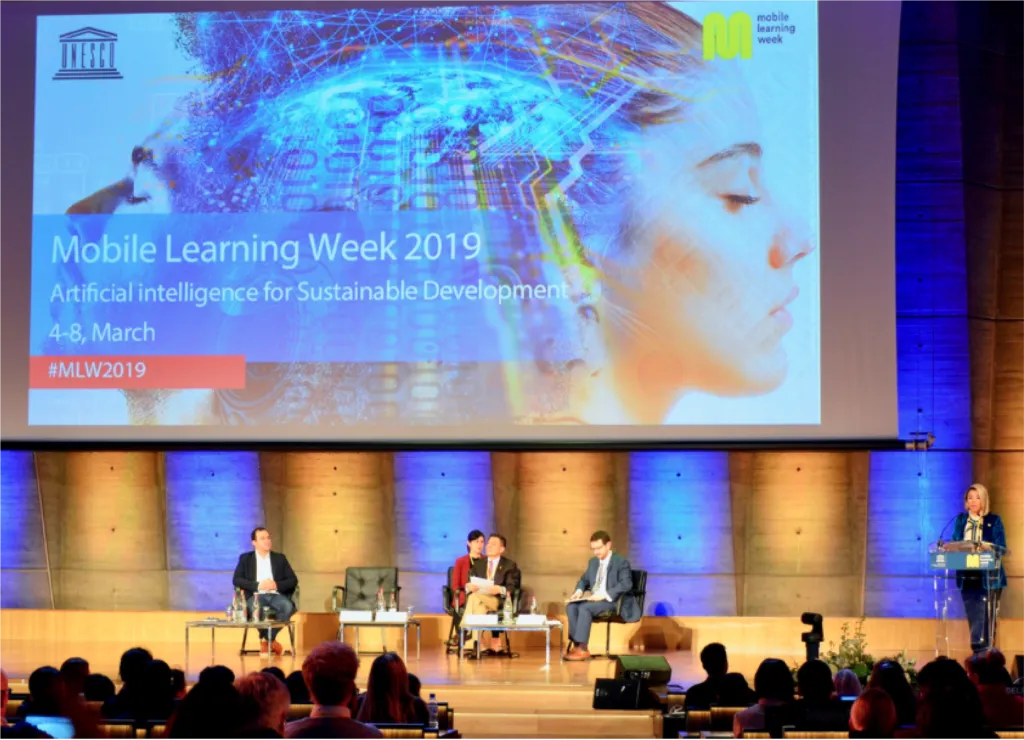 Photo of the stage with professional speakers seated and standing below a large format screen at the UNESCO Mobile Learning Week 2019