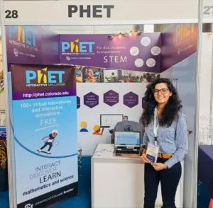 A woman in glasses with black hair stands in front of a conference booth. The booth features a standing banner, laptop, and other materials advertising PhET Interactive Simulations.