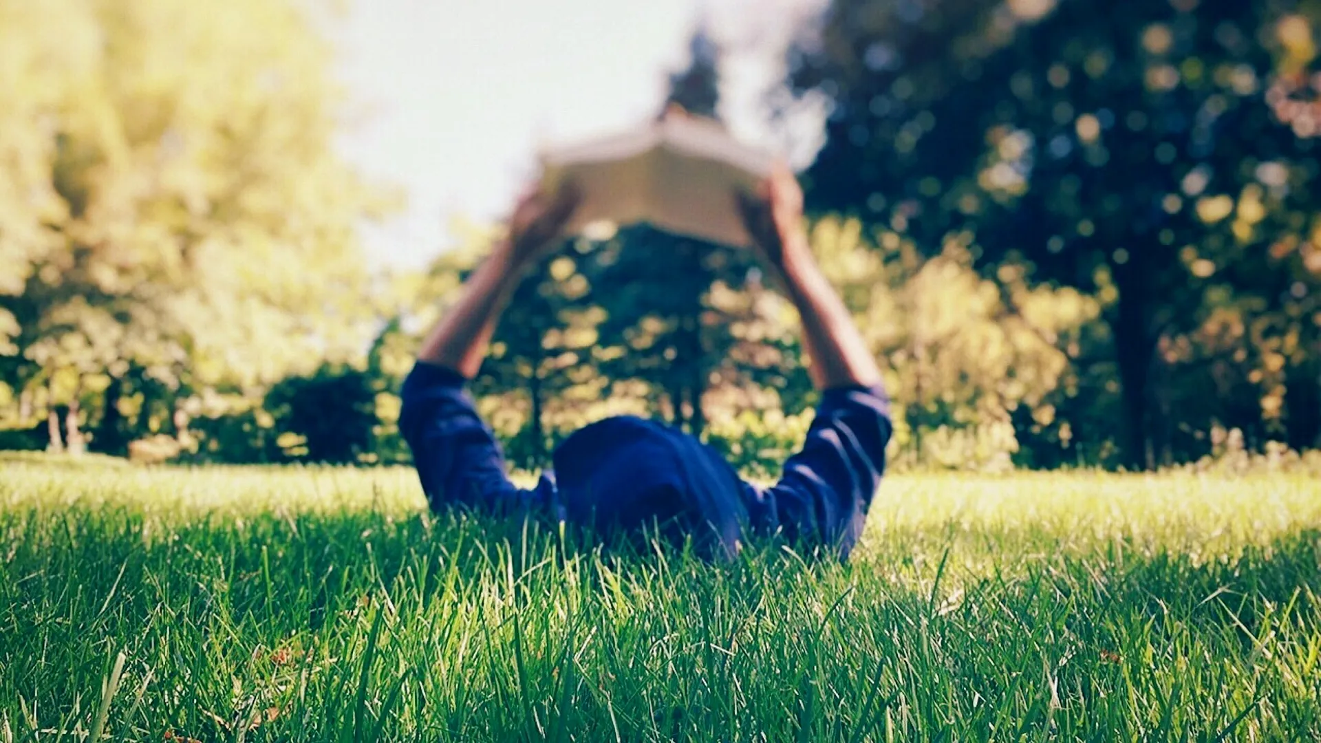 A person lying on their back in the grass on a sunny day, reading a book.