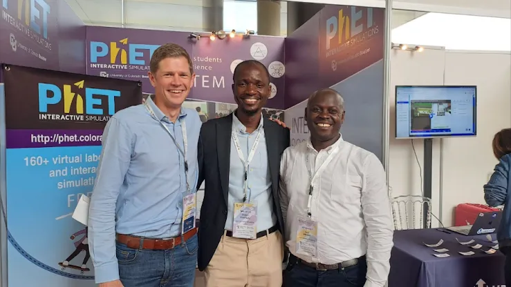 Three men posing together for a picture in front of the PhET exhibition booth.