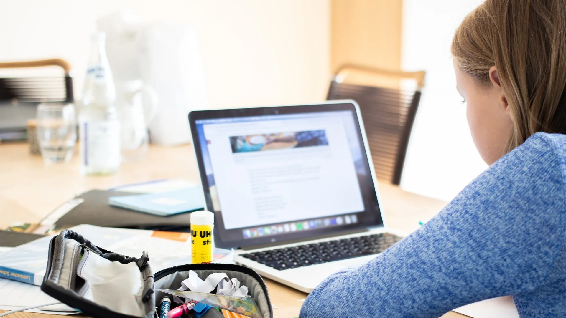 A girl sitting at a table and using a laptop. A pencil case and other classroom materials sit on the table next to her.