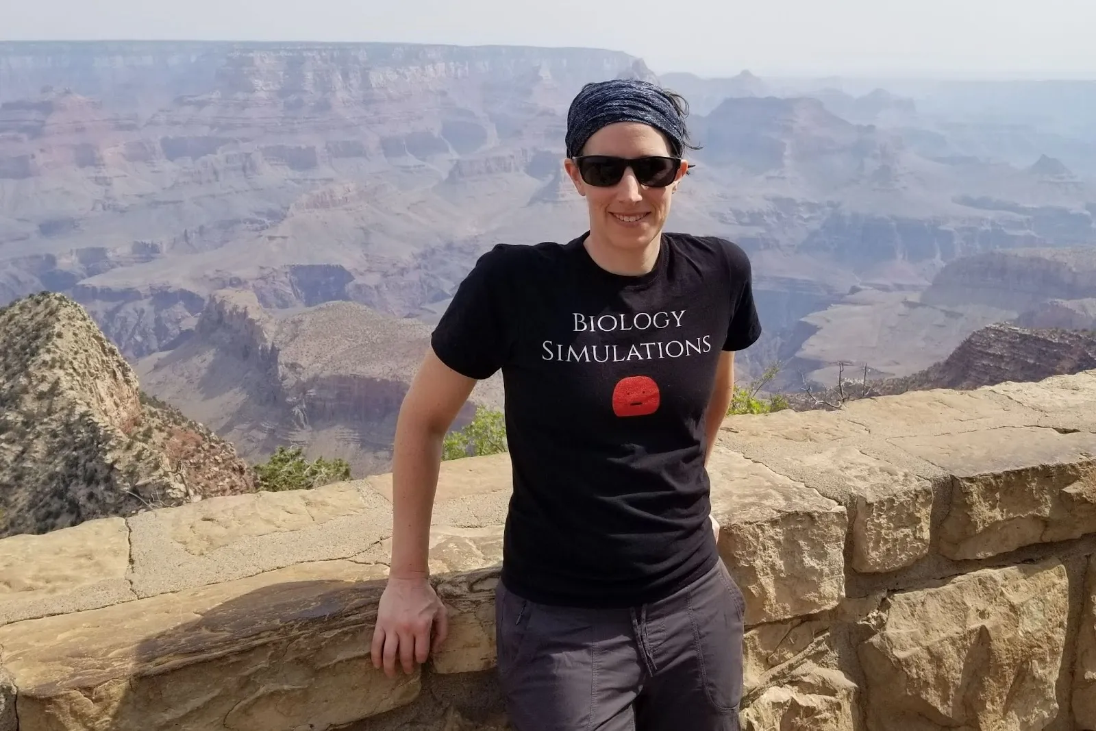 A woman, Jolene Pappas, in sunglasses and a t-shirt that reads "Biology Simulations" stands at the edge of the Grand Canyon.