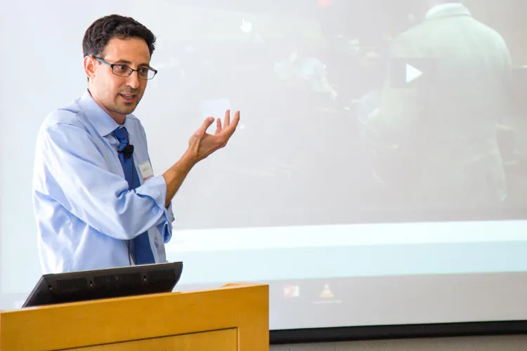 Josh Bookin, a man with dark hair, glasses, a light blue shirt, and dark blue tie, stands at a podium and gestures to a projection screen behind him.