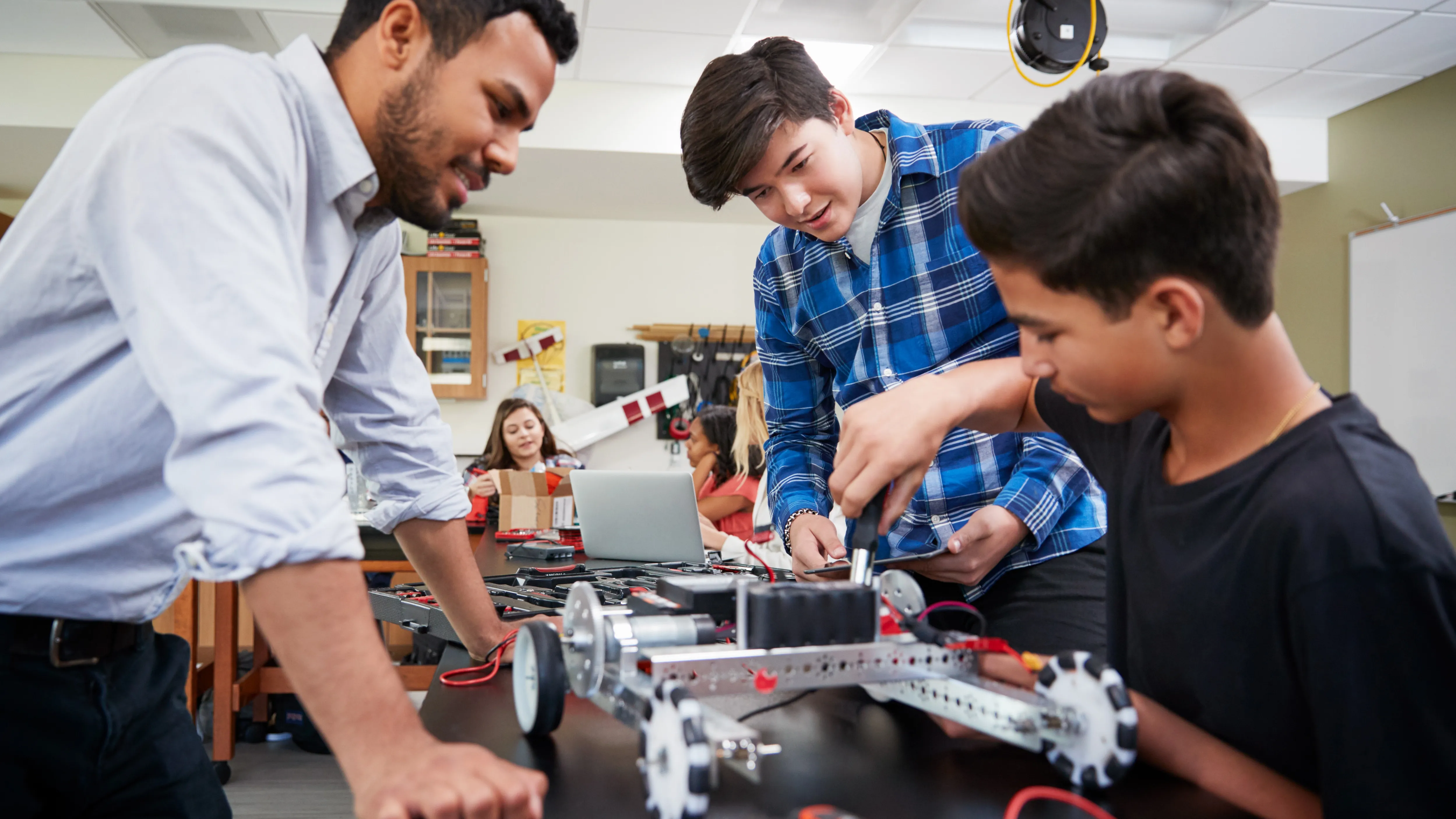 Two students work on building a robot in a classroom, as a teacher watches over them, assisting.