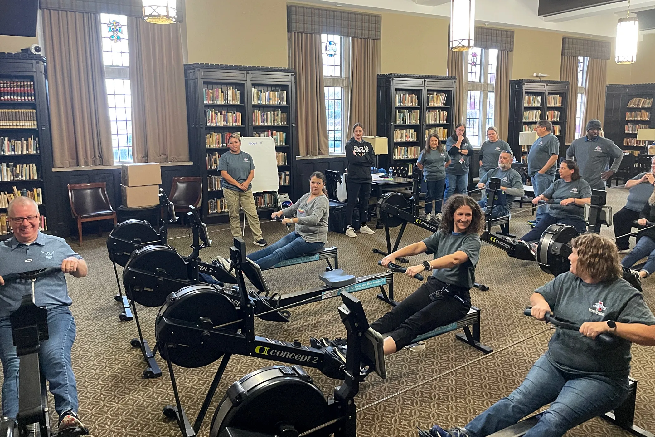 Rowing coach Neil Bergenroth demonstrates the correct form on a rowing machine while a group of adult learners follow along on their own machines, with other learners waiting and observing in the background. They are all smiling and enjoying the experience.