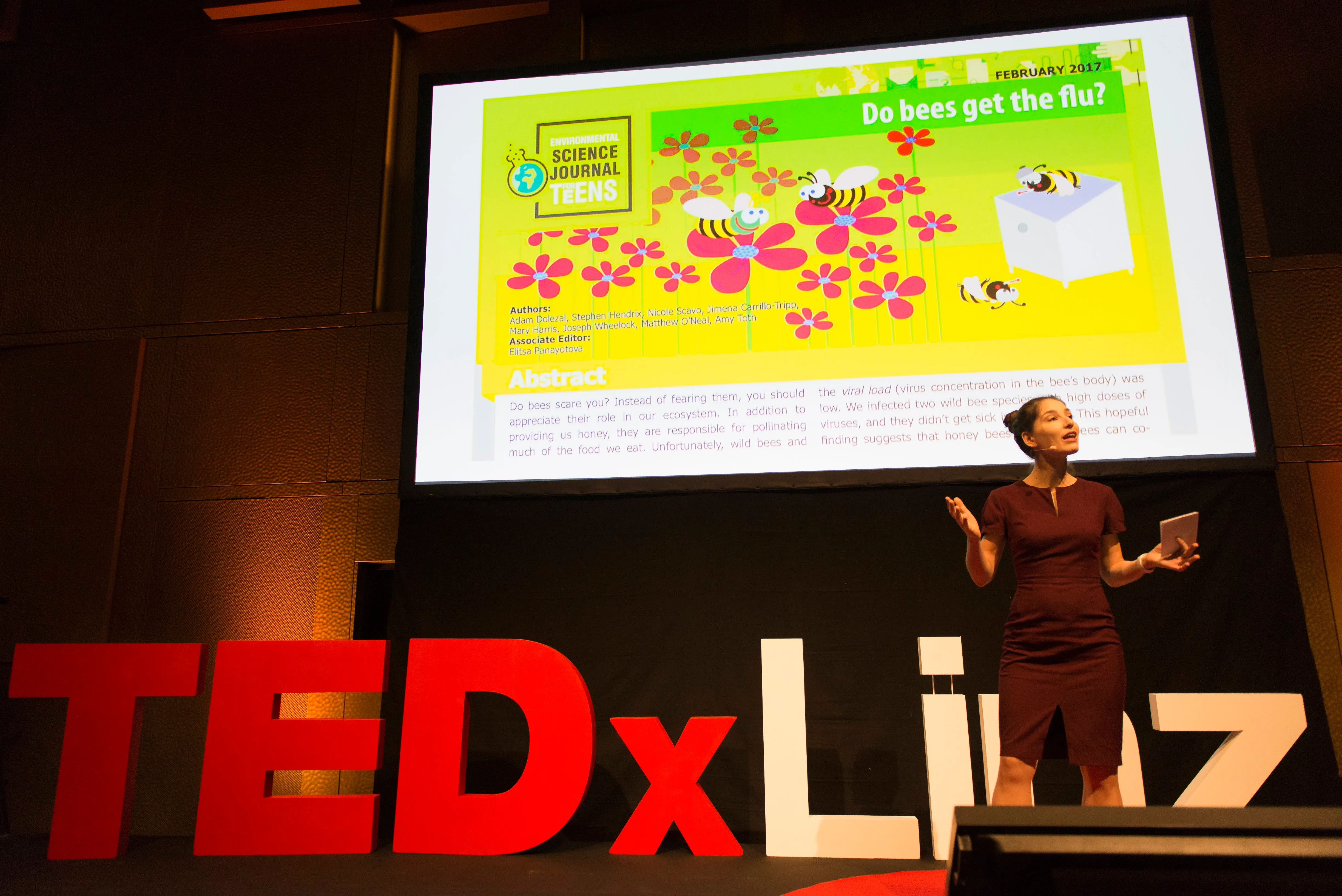Tanya Dimitrova, a woman with dark brown hair in a bun and wearing a brown dress, stands on a stage at a TEDx event. Behind her is a slide about a question ("Do bees get the flu?") which was published in the Science Journal for Kids.
