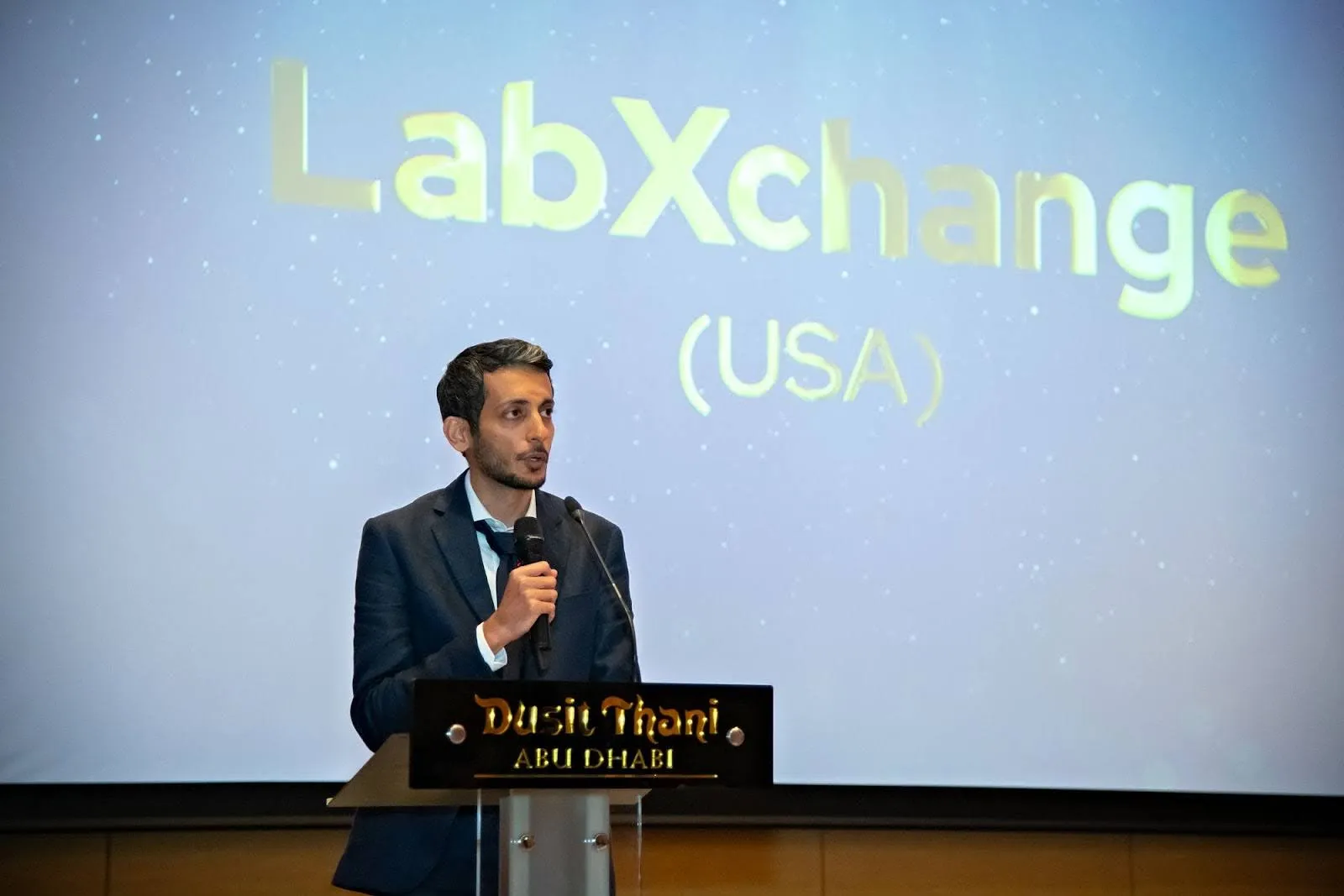 Usman Khalid, Chief Technology Officer at LabXchange, speaks at the QS Reimagine Education Awards ceremony. He is a tan-skinned man with dark hair wearing a dark blue suit and standing on a stage in front of podium, with a microphone in his hand. In the background, a projection screen says "LabXchange (USA)".