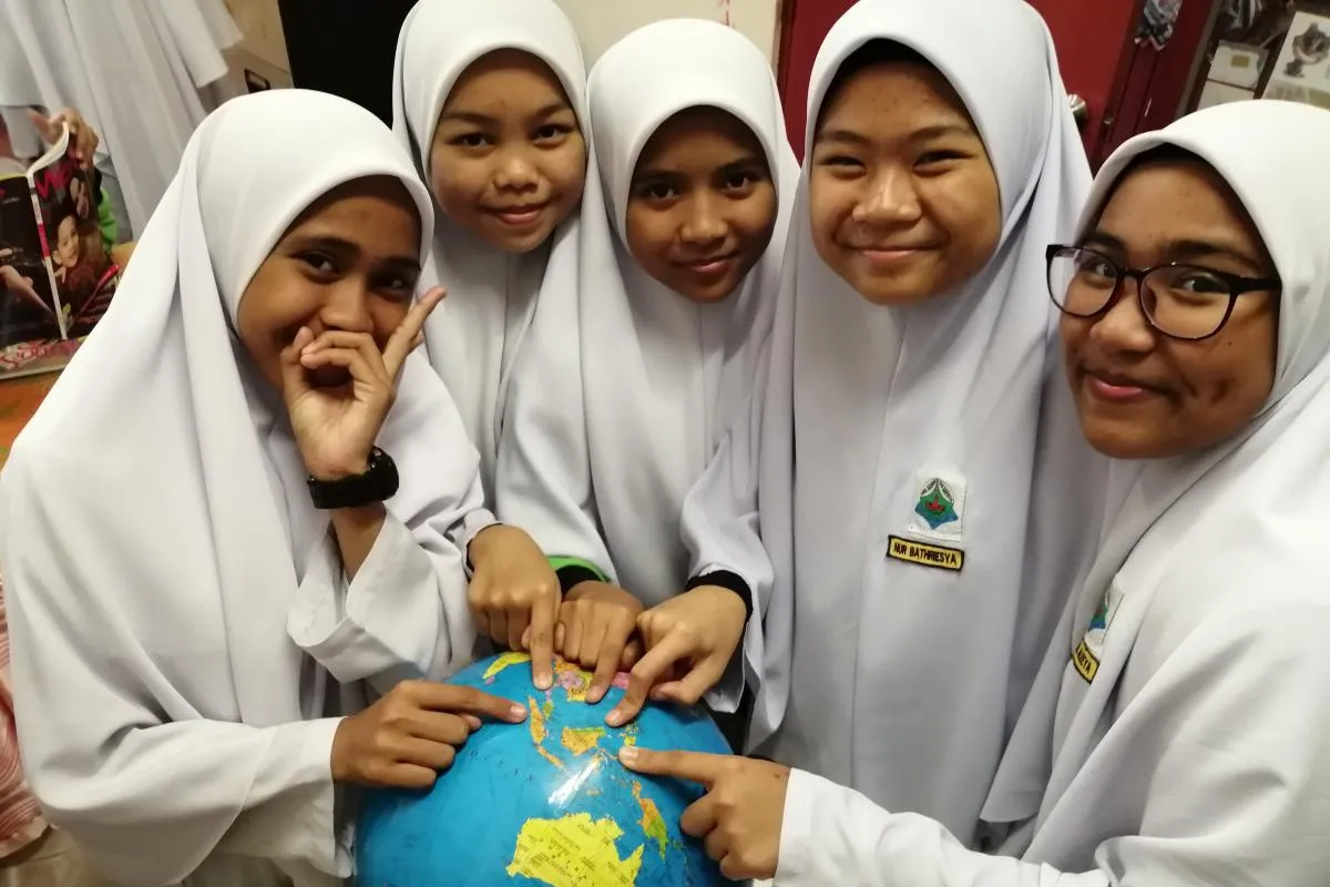 Five young students wearing white Indonesian tudong-style hijabs smile at the camera while pointing to Indonesia on a globe in front of them.