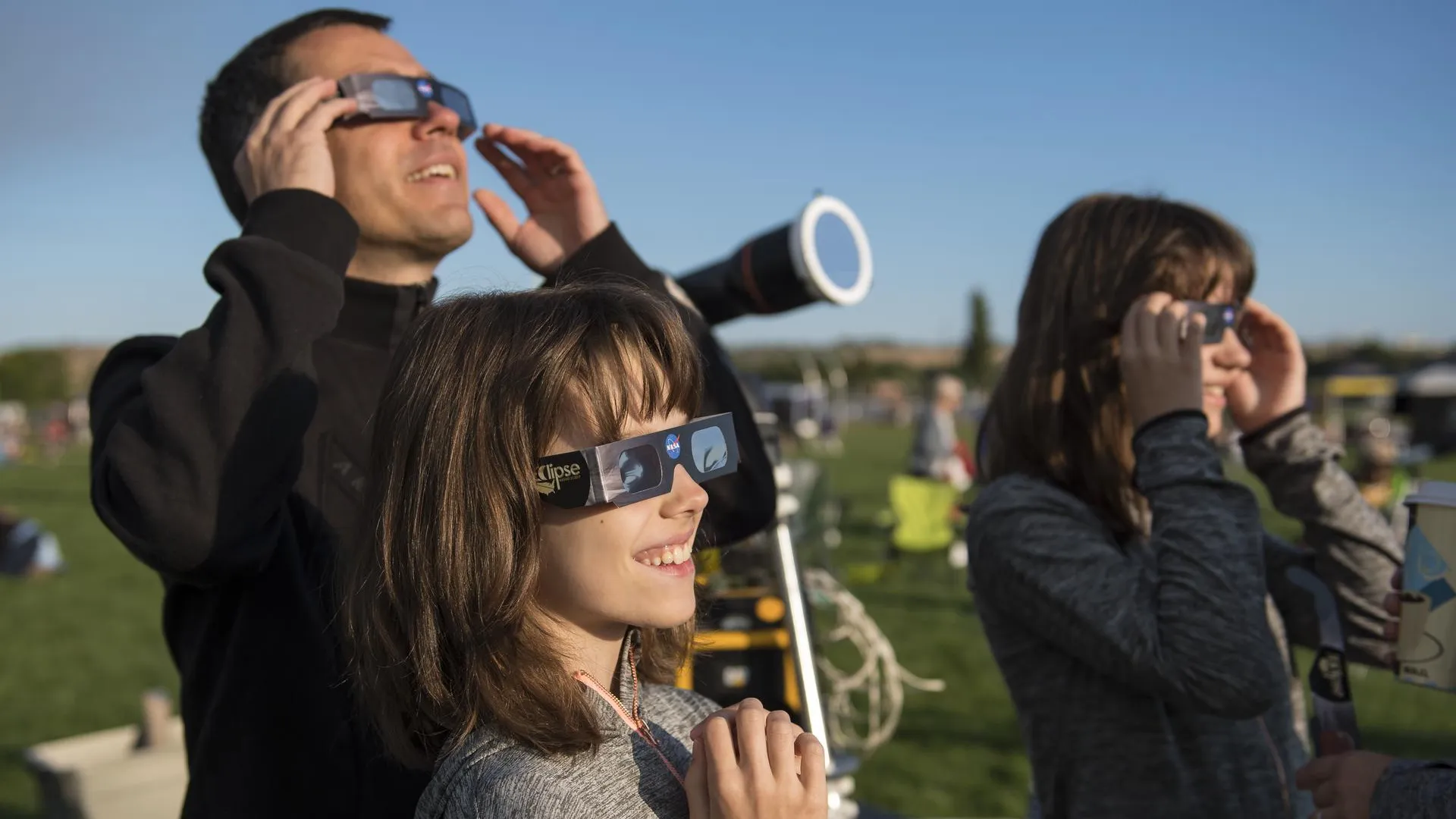 A adult man and two teenagers observe the 2017 total solar eclipse in Oregon, USA. They are all wearing eclipse glasses and gazing up at the sky. Photo by NASA/Aubrey Gemignani.