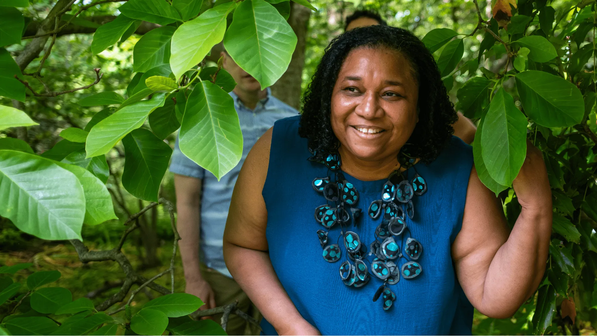 A photograph of Dr. Catherine Quinlan and her family, standing under the leafy branches of a tree. Dr. Quinlan (a Black woman with shoulder-length curly hair wearing a blue blouse and a long necklace) is in the forefront of the photograph, smiling cheerfully at the camera. Her husband (a white man) and her son are standing behind her, their features obscured by the foliage.