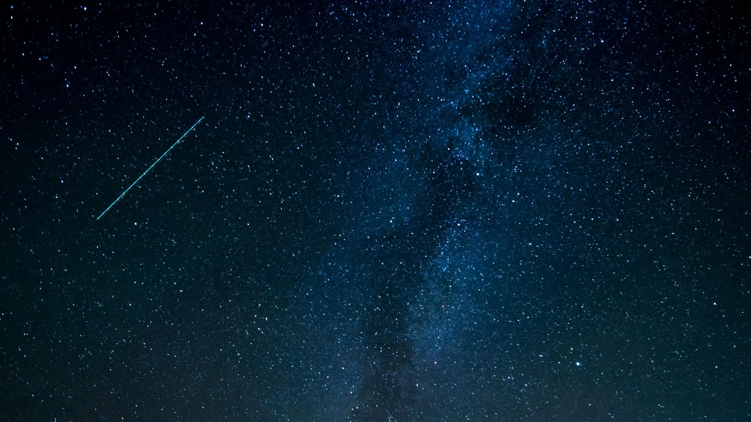 Against a dark-blue and black night sky, there are thousands of bright stars, as well as the glow of the Milky Way. In the upper left corner of the image, a meteor streaks across the sky.