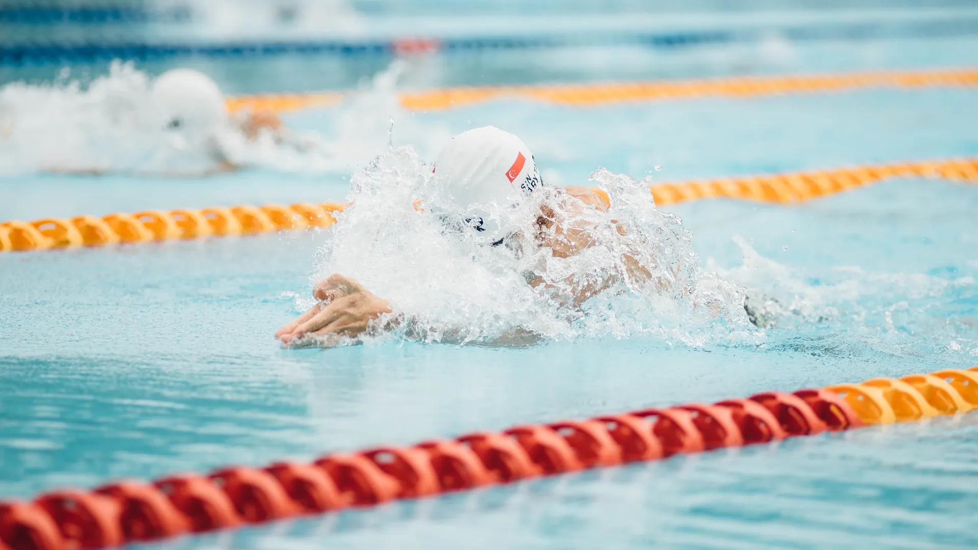 In the foreground, a swimmer swims intensely in the lane of a swimming pool during a race. Another swimmer can be seen in a separate lane in the background.