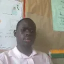 A Black man with short black hair in a pink shirt, sitting in a classroom.