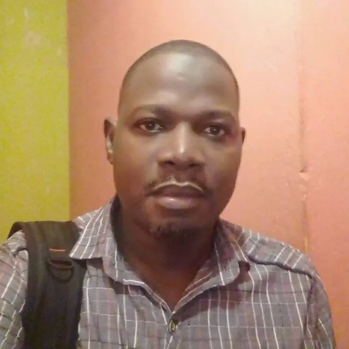 A Black man with short black hair and a goatee, wearing a grey dress shirt and carrying a backpack over his right shoulder.