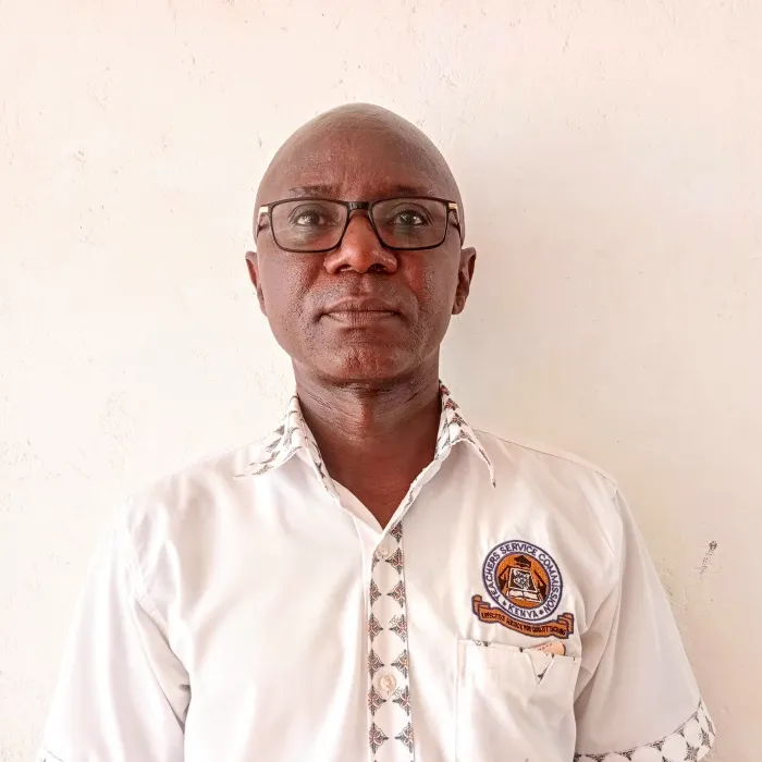 A Black man with glasses and a white shirt with patterning along the collar and hems standing against a white wall.