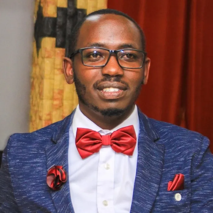 A Black man with short black hair and a short beard, wearing a blue blazer, white dress shirt, and red bowtie, with a red handkerchief in his blazer pocket and red flower pinned to his lapel. 