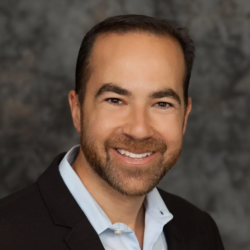 A white man with short dark hair and a short beard wearing a black blazer and blue collared shirt.