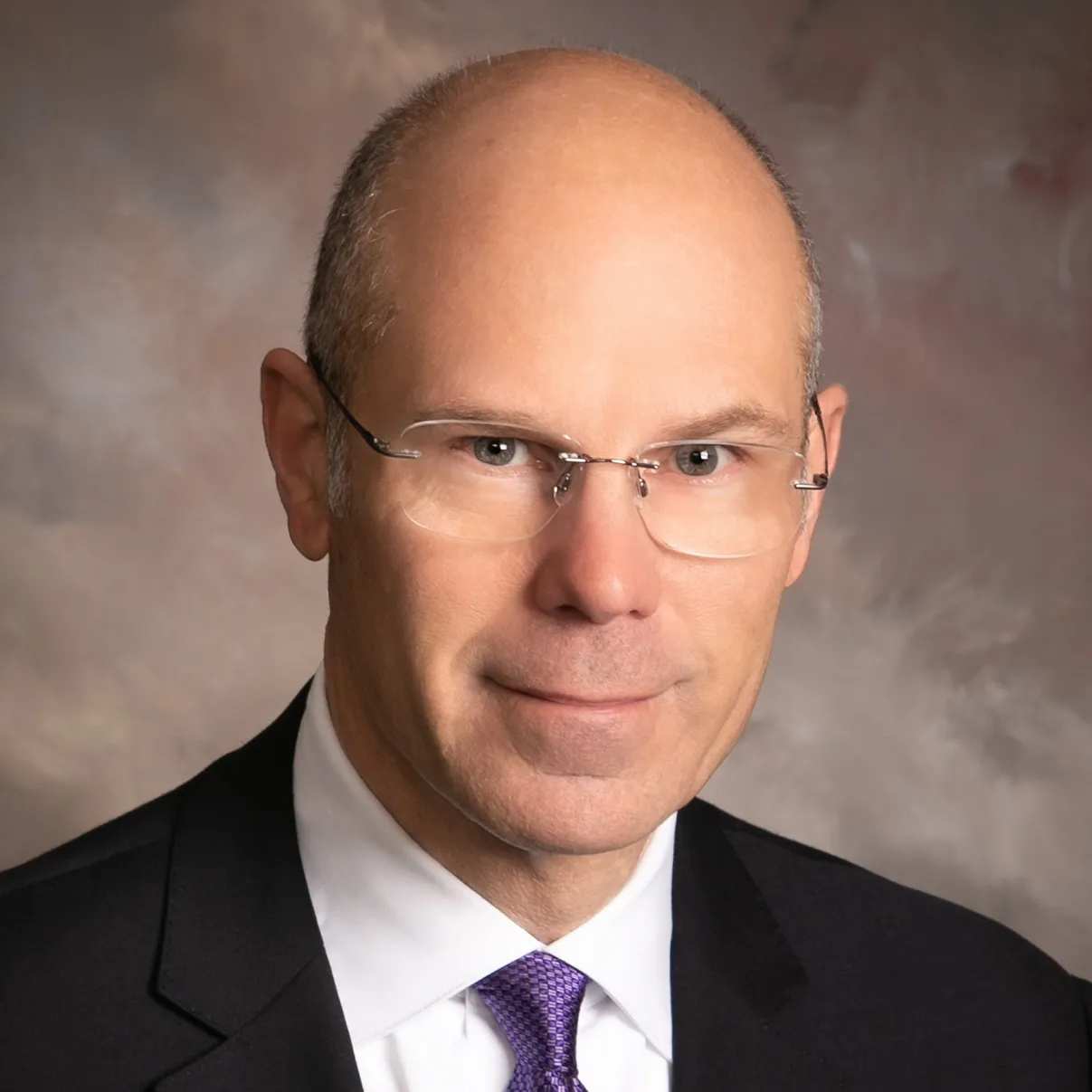 A white man with glasses wearing a black suit and tie.