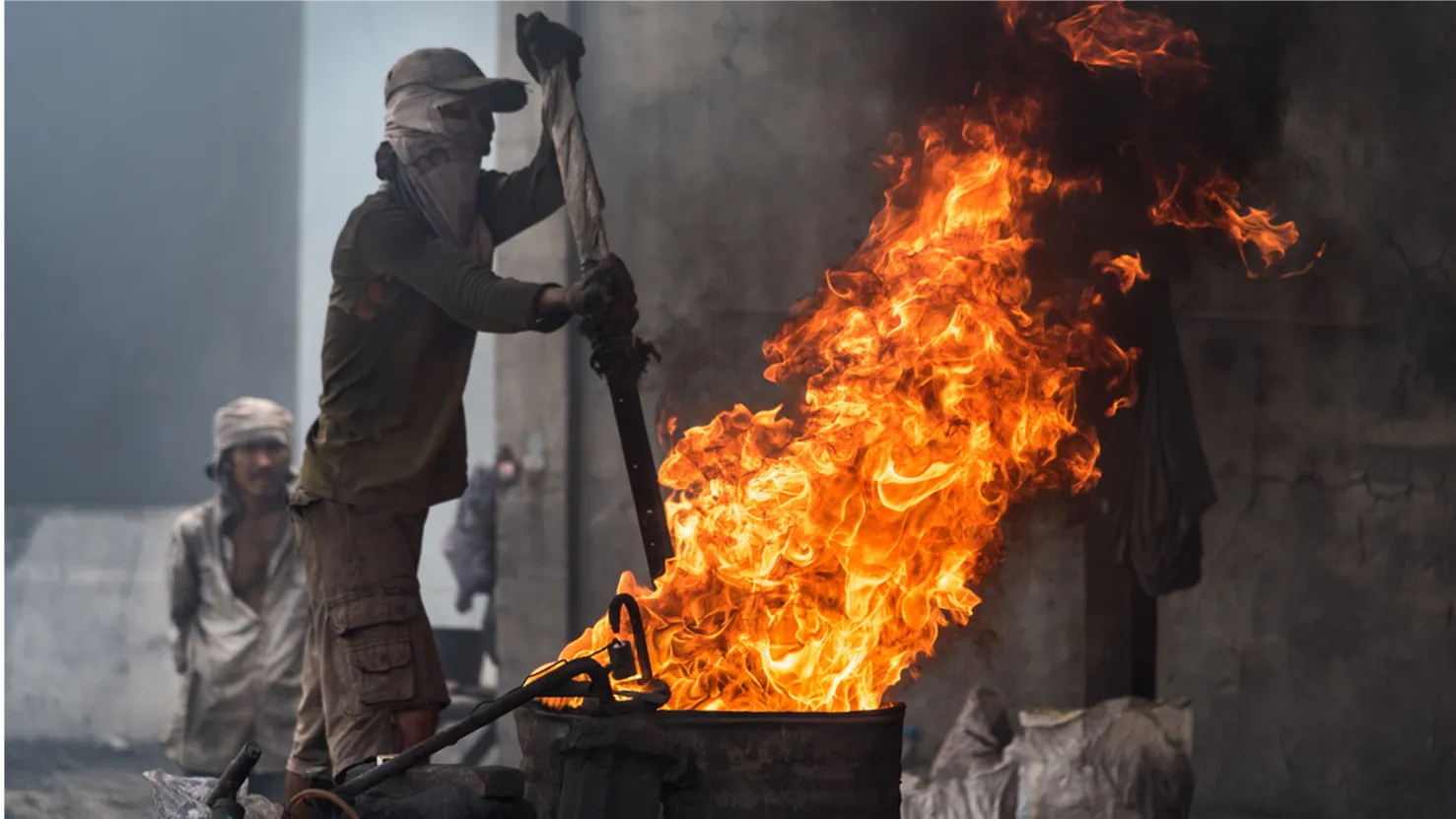 A man wearing work clothes and a cloth face covering carefully pokes the inside of a flaming barrel with a large metal post.