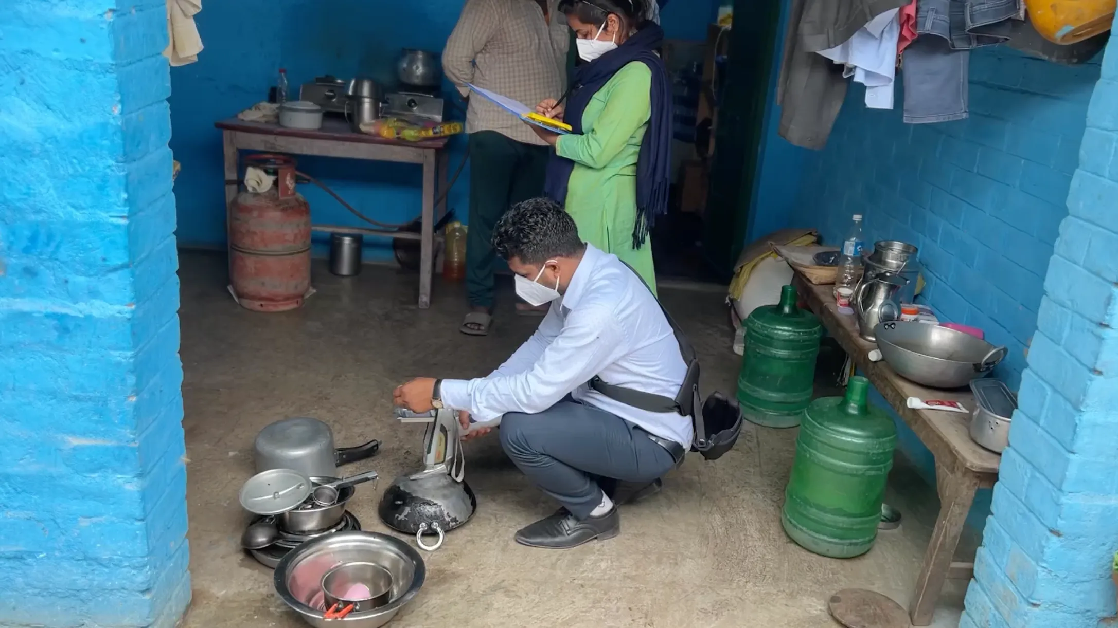 In the foreground, a man crouches over a pile of aluminum pots, testing them for lead using a digital meter. In the background, a woman records data on a clipboard.