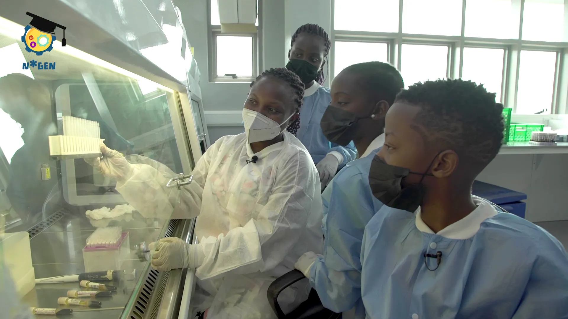 A group of kids wearing PPE look at a series of test tubes under a fume hood.
