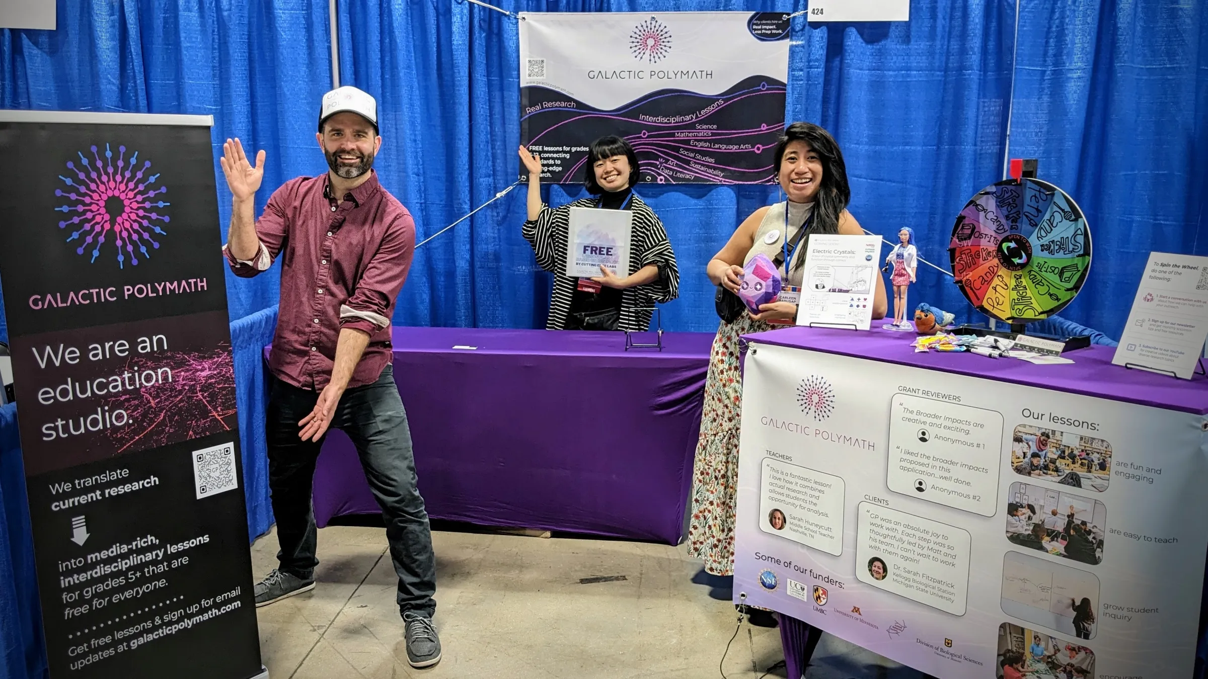 The Galactic Polymath team highlighting their free learning materials at a convention. A man stands on the left side of the image next to a banner displaying information about Galactic Polymath. On the right, a woman is leaning against a tall table with a "wheel of fortune" on it, used for giving away swag. In the background, a woman stands behind a low table and points at a banner above her that reads "Galactic Polymath." 