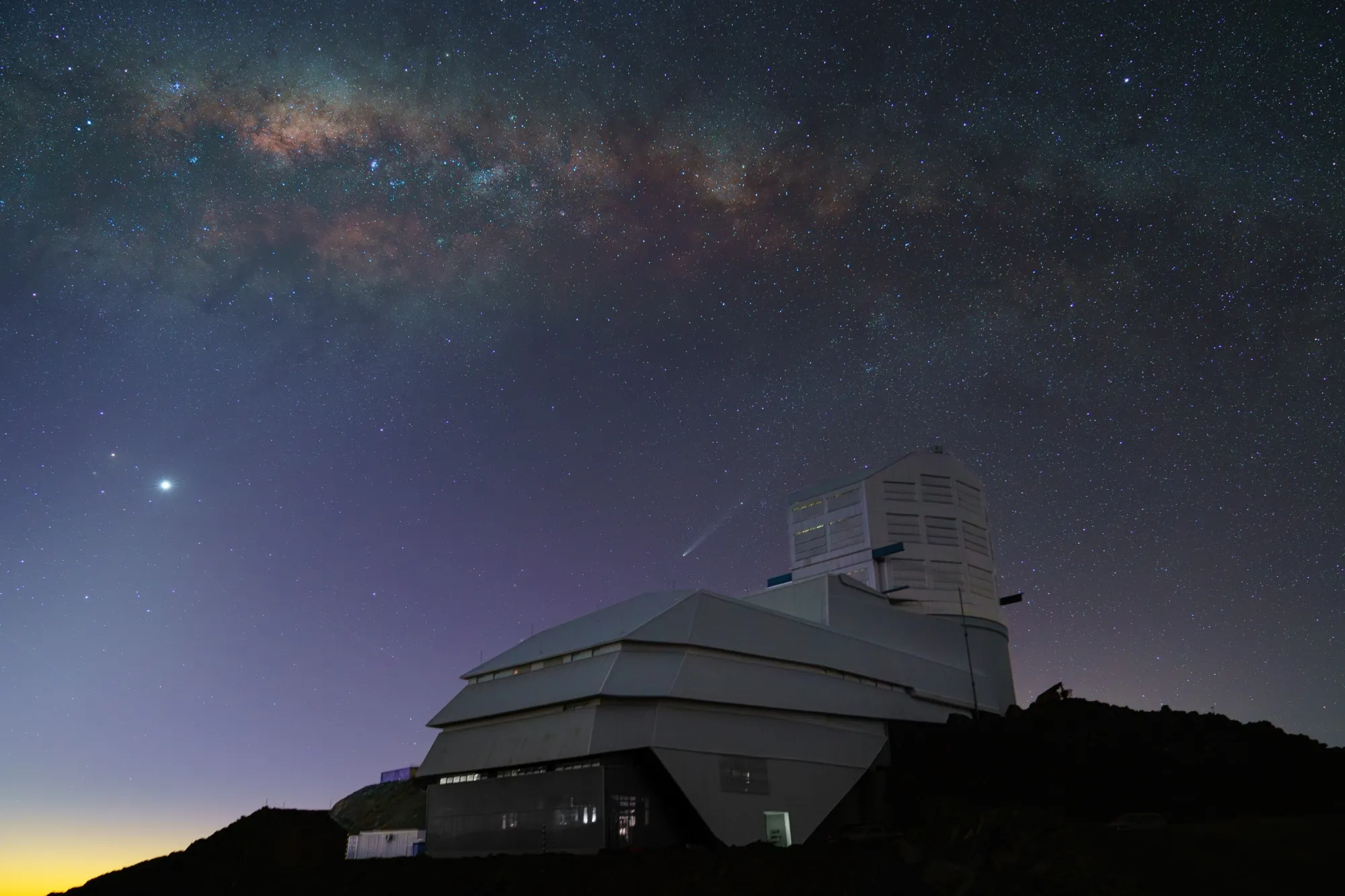 The Vera C. Rubin Observatory in Chile, located on a mountain peak. In the background is a long exposure of the night sky, showing the Milky Way galaxy and various stars. Credit: RubinObs/NOIRLab/SLAC/NSF/DOE/AURA.