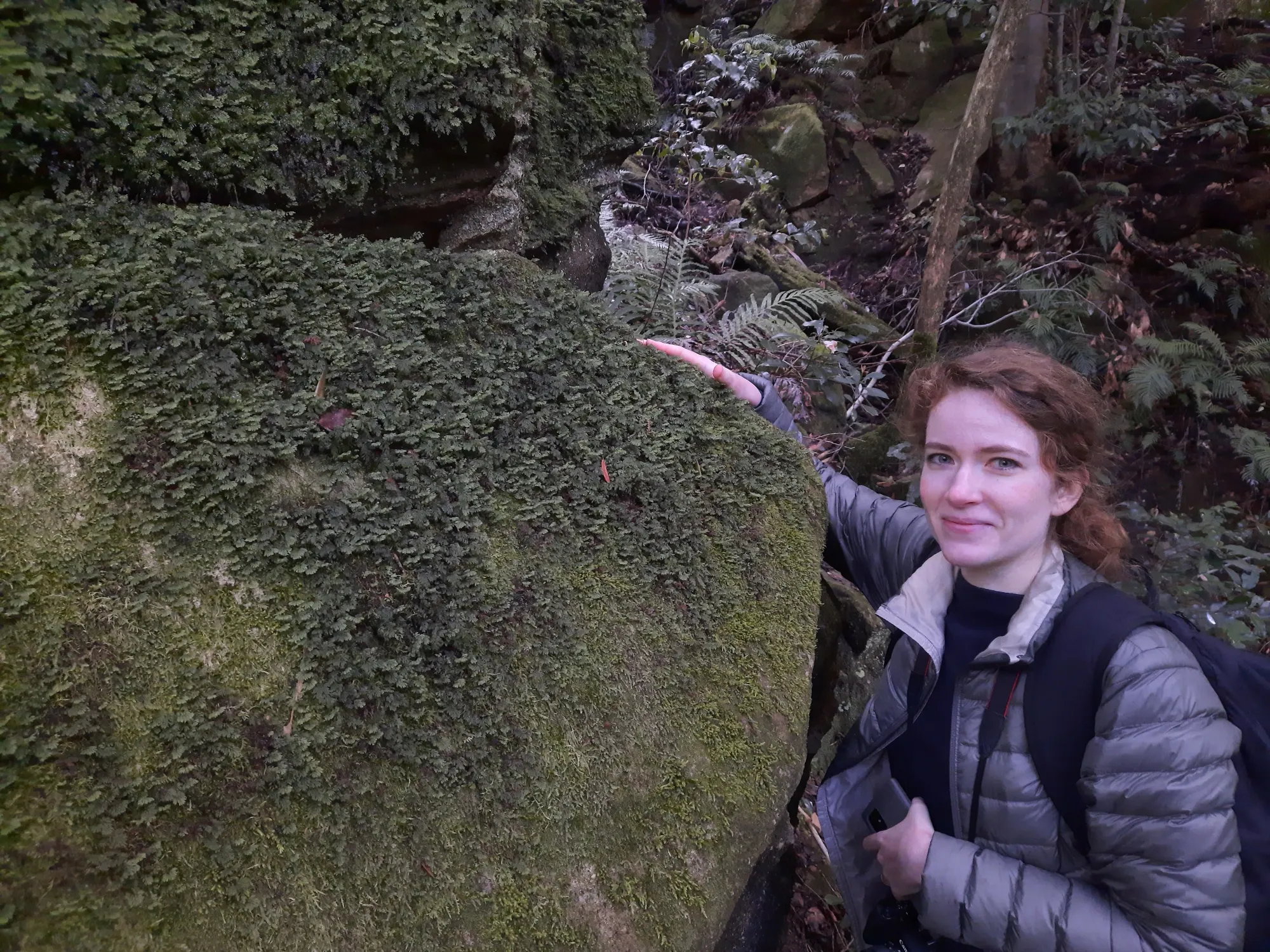 A woman stands in a forest next to a large rock that has moss and ferns growing on it.